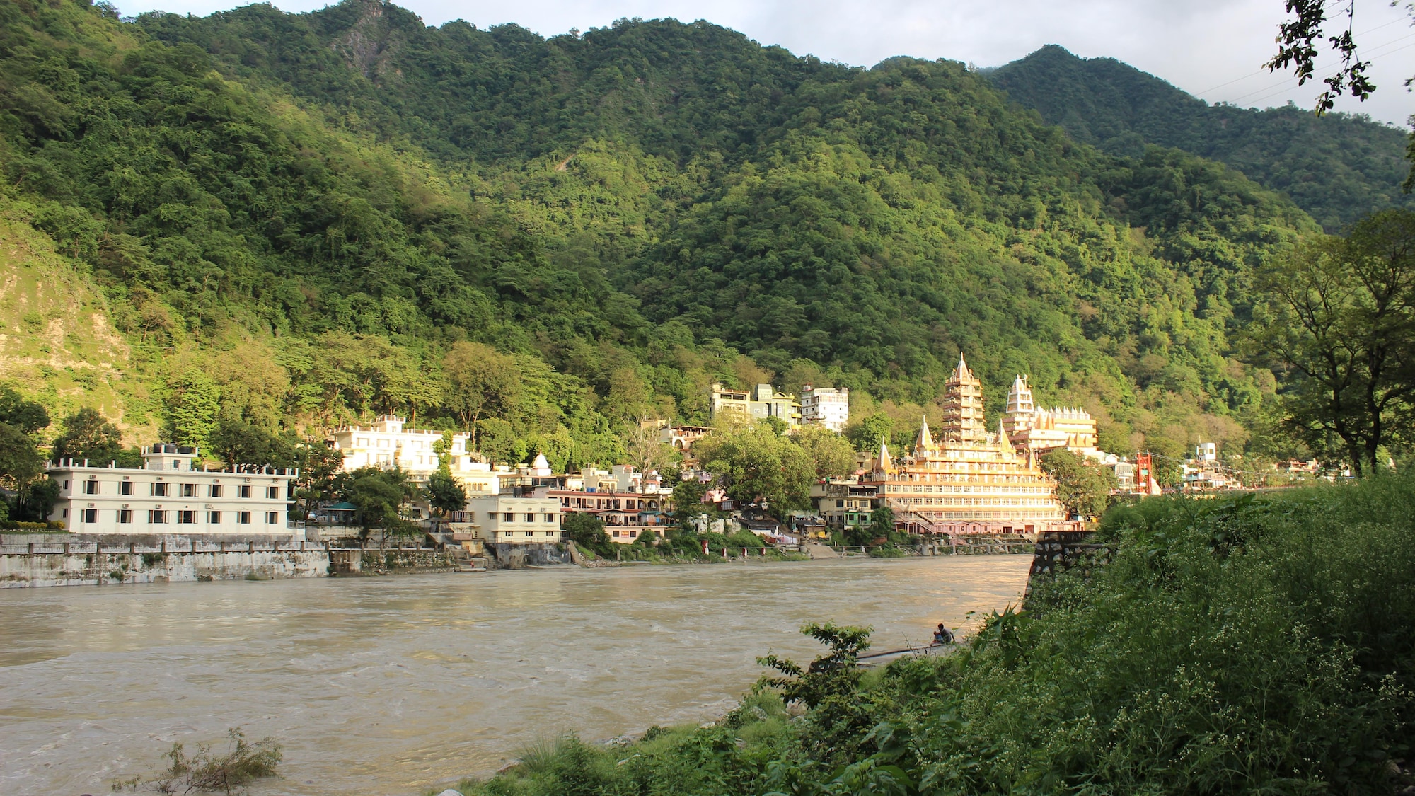 A river flows past buildings with tree-covered hills in the background.