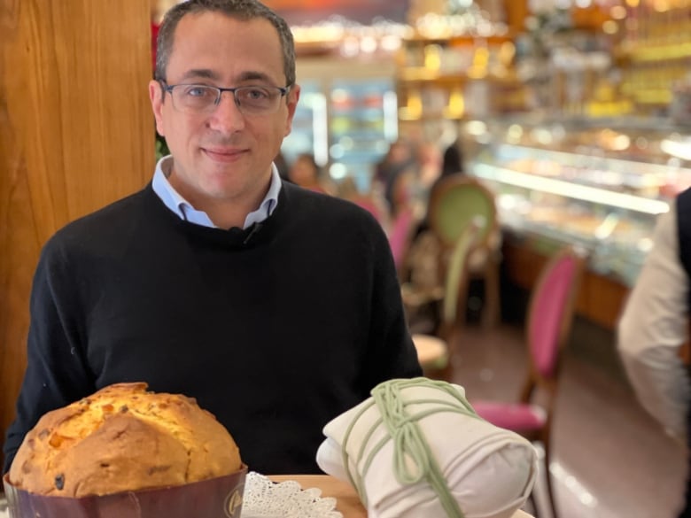 A smiling man wearing a sweater and glasses is pictured at a table with a sweet baked bread and a white cloth pouch tied with a green string.